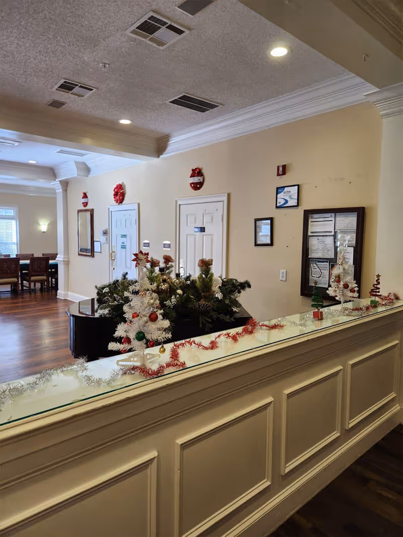 Interior view of a senior living facility lobby or common area decorated for the holidays with small white Christmas trees, garlands, and festive ornaments on a glass-topped counter. The room has beige walls, white doors, framed notices on the wall, and wooden flooring. In the background, there is a dining area with tables and chairs.