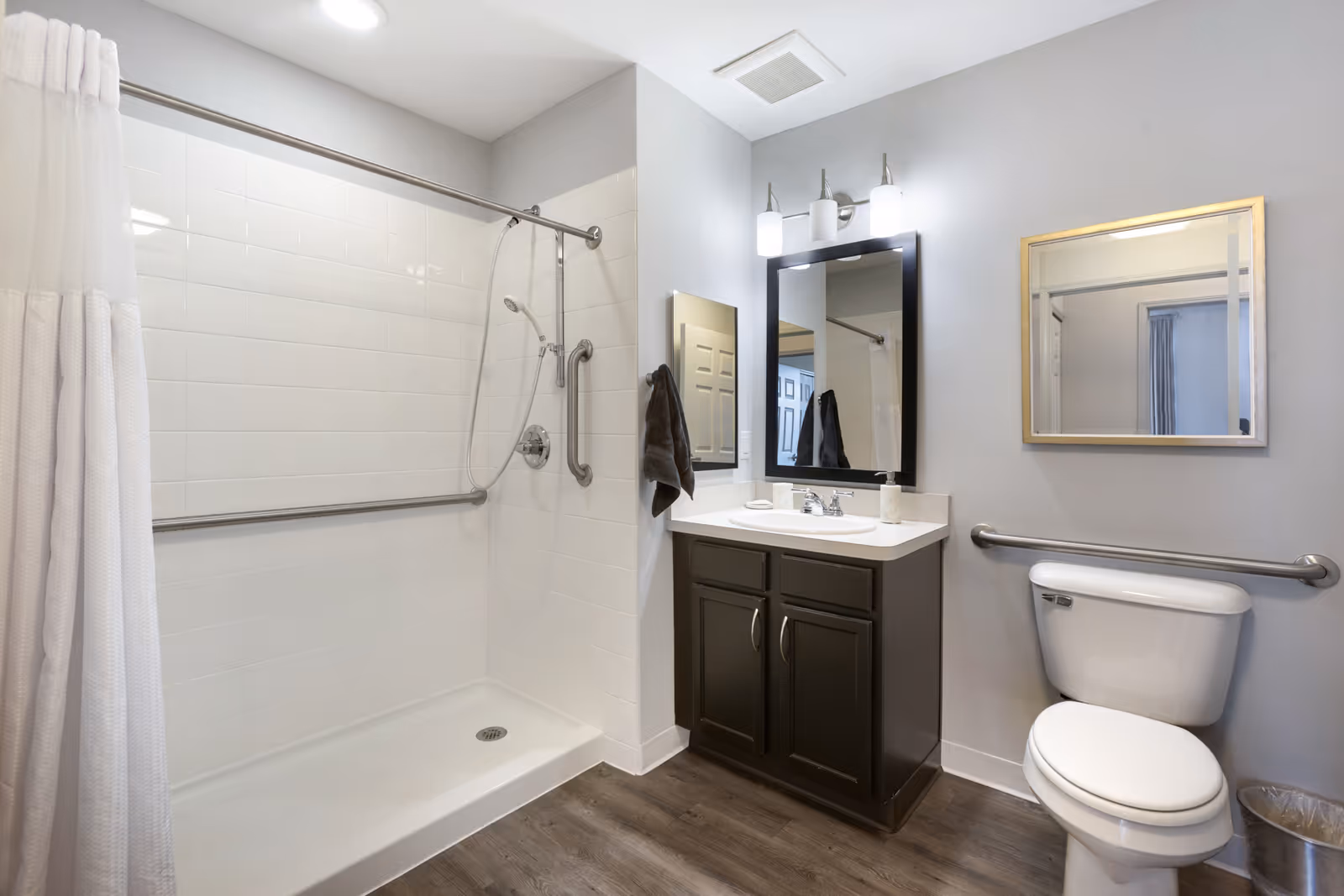 Accessible bathroom with a walk-in tiled shower, vanity and mirror, and a toilet with grab bars.