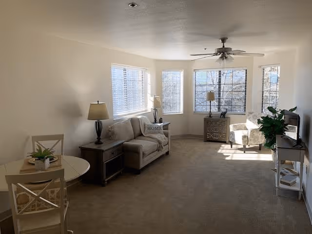 A bright living room with beige carpet and white walls featuring a small round dining table with two chairs, a beige loveseat with a 'HOME' pillow, two side tables with lamps, a patterned armchair near a large window, a ceiling fan, a TV on a stand, and a green potted plant.