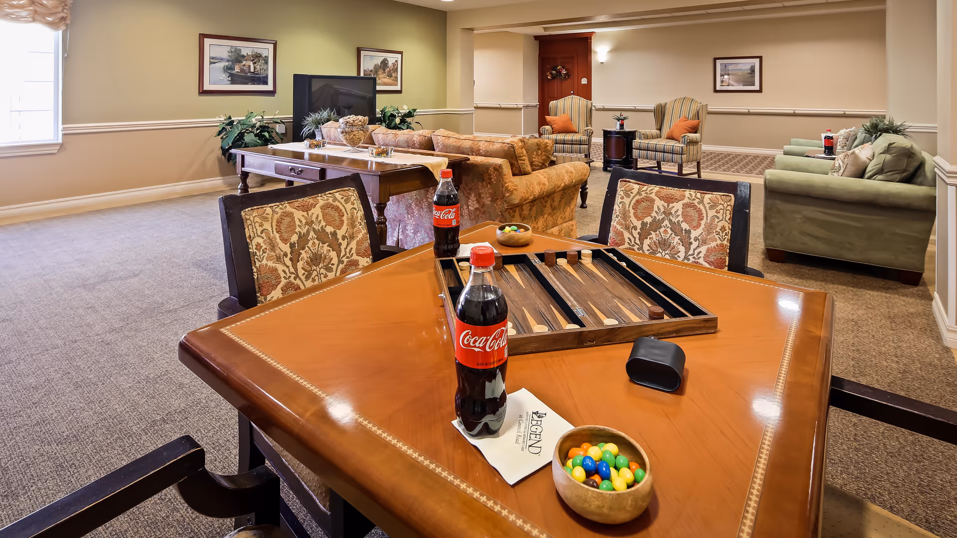 A cozy common area in a senior living facility featuring a wooden table with a backgammon game, two bottles of Coca-Cola, a bowl of colorful candies, and patterned chairs. In the background, there are several upholstered sofas and armchairs arranged for socializing, with framed artwork on the walls and a window letting in natural light.