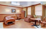 A spacious senior living bedroom with a single bed covered in a patterned bedspread, a wooden chair, a small table with a flower arrangement, a comfortable armchair, and large windows with blinds and curtains allowing natural light to fill the room.