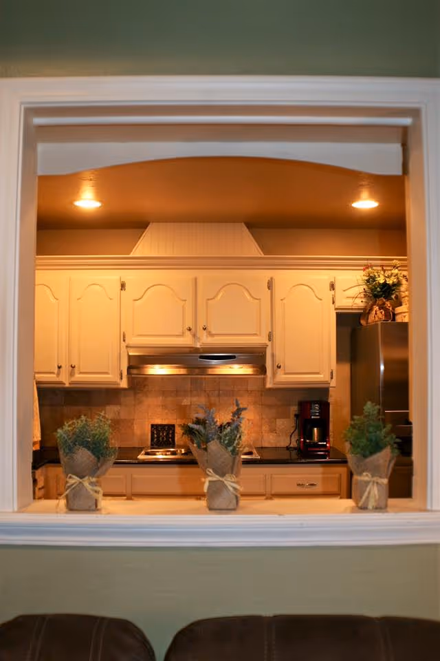 View through a pass-through window into a kitchen with cream-colored cabinets, a stainless steel range hood, a coffee maker on the counter, and three small potted plants wrapped in burlap on the window ledge.