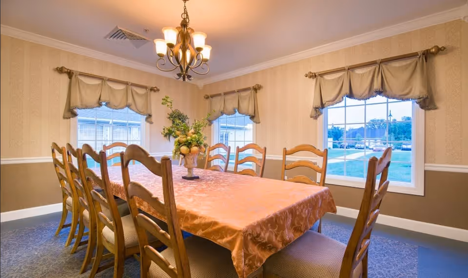A dining room with a long rectangular table covered with a peach-colored tablecloth. The table is surrounded by wooden chairs with cushioned seats. There is a floral centerpiece on the table. The room has three large windows with beige valances, beige patterned wallpaper, and a chandelier hanging from the ceiling. Outside the windows, a grassy area and other buildings are visible.