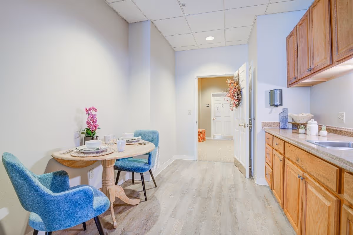 A small dining area with a round wooden table set for two, featuring blue upholstered chairs and a pink orchid centerpiece. To the right is a kitchen counter with wooden cabinets, a sink, and various kitchen items. The room has light-colored walls and flooring, with a doorway leading to another room in the background.