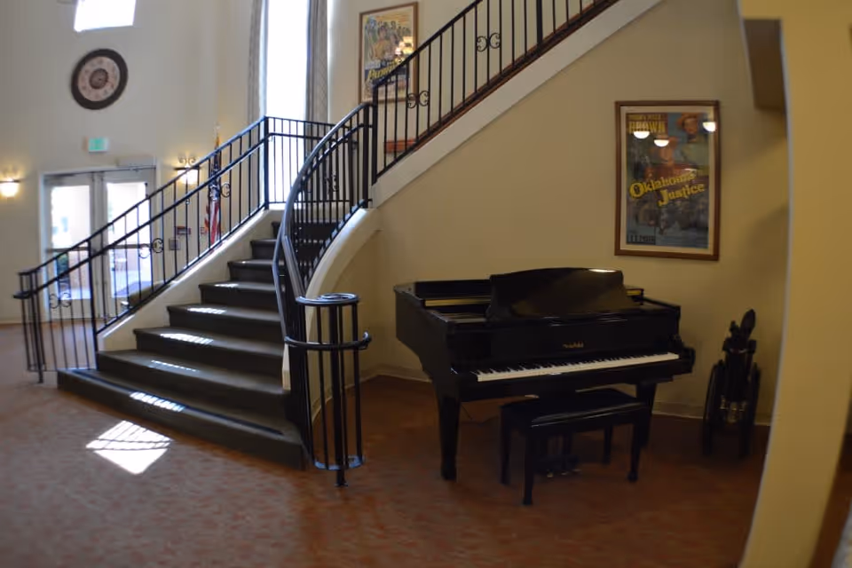 Interior view of a senior living facility showing a curved staircase with black railings, a black grand piano with a matching bench, a wheelchair beside the piano, and framed posters on the walls. The area is carpeted and well-lit with natural light coming through tall windows near the entrance door.