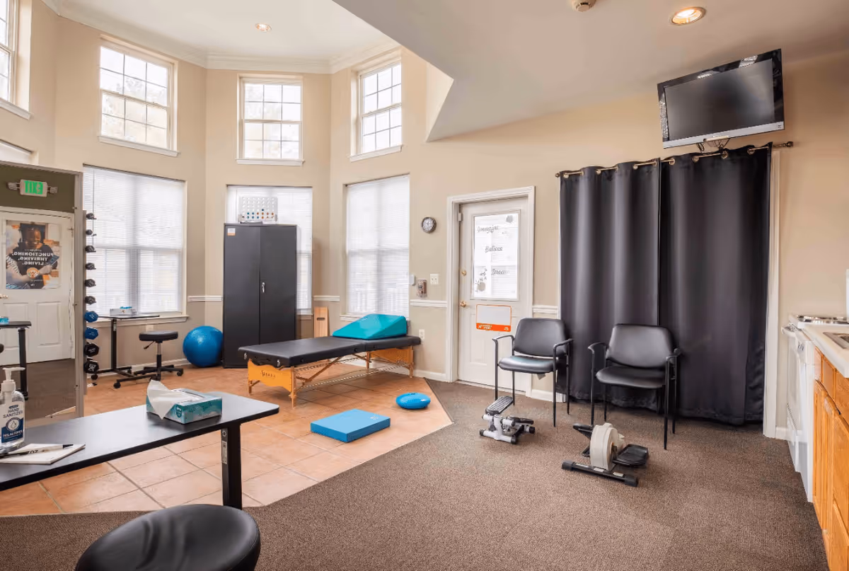 A bright therapy or exercise room with large windows, a black therapy table, exercise equipment including a blue exercise ball, weights, and a small pedal exerciser. There are two black chairs, a wall-mounted TV, a black curtain, and a door with motivational words on the glass. The room has a mix of tiled and carpeted flooring.