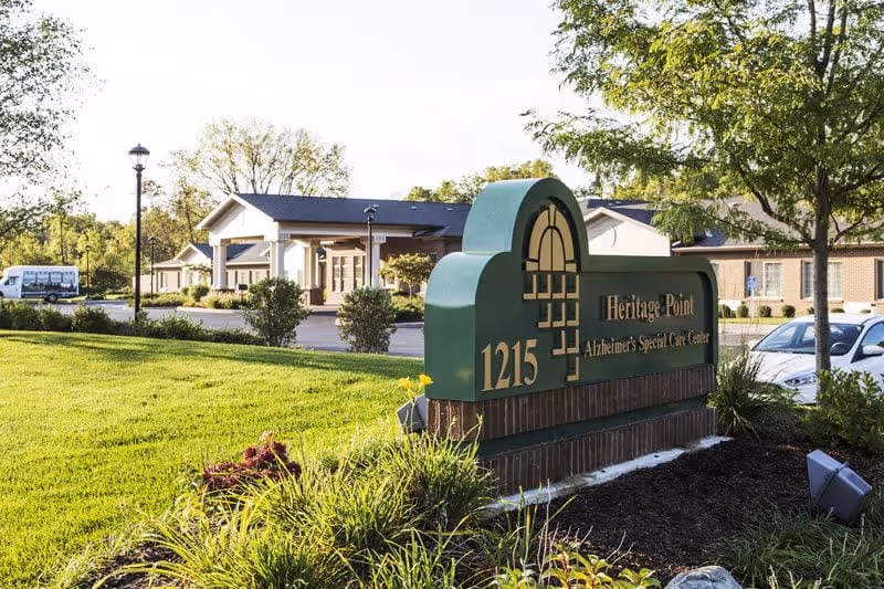 A landscaped lawn and sign reading "Heritage Point Alzheimer's Special Care Center 1215" with the facility building and parked cars visible behind it.