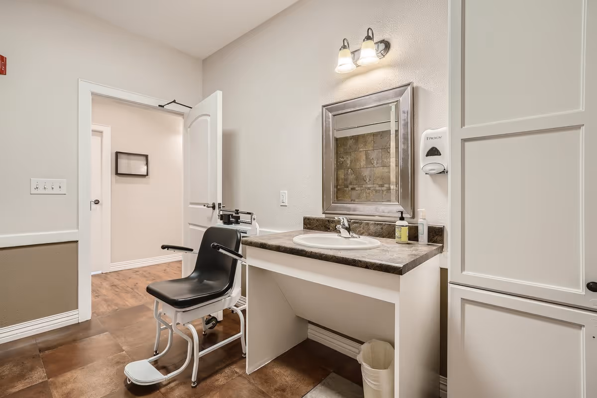 A clean and well-lit bathroom area in a senior living facility featuring a sink with a marble countertop, a large framed mirror above it, a soap dispenser mounted on the wall, and a black shower chair with armrests positioned near the sink. The floor has brown tiles, and the walls are painted in neutral tones. The door to the room is open, showing a hallway with wooden flooring.