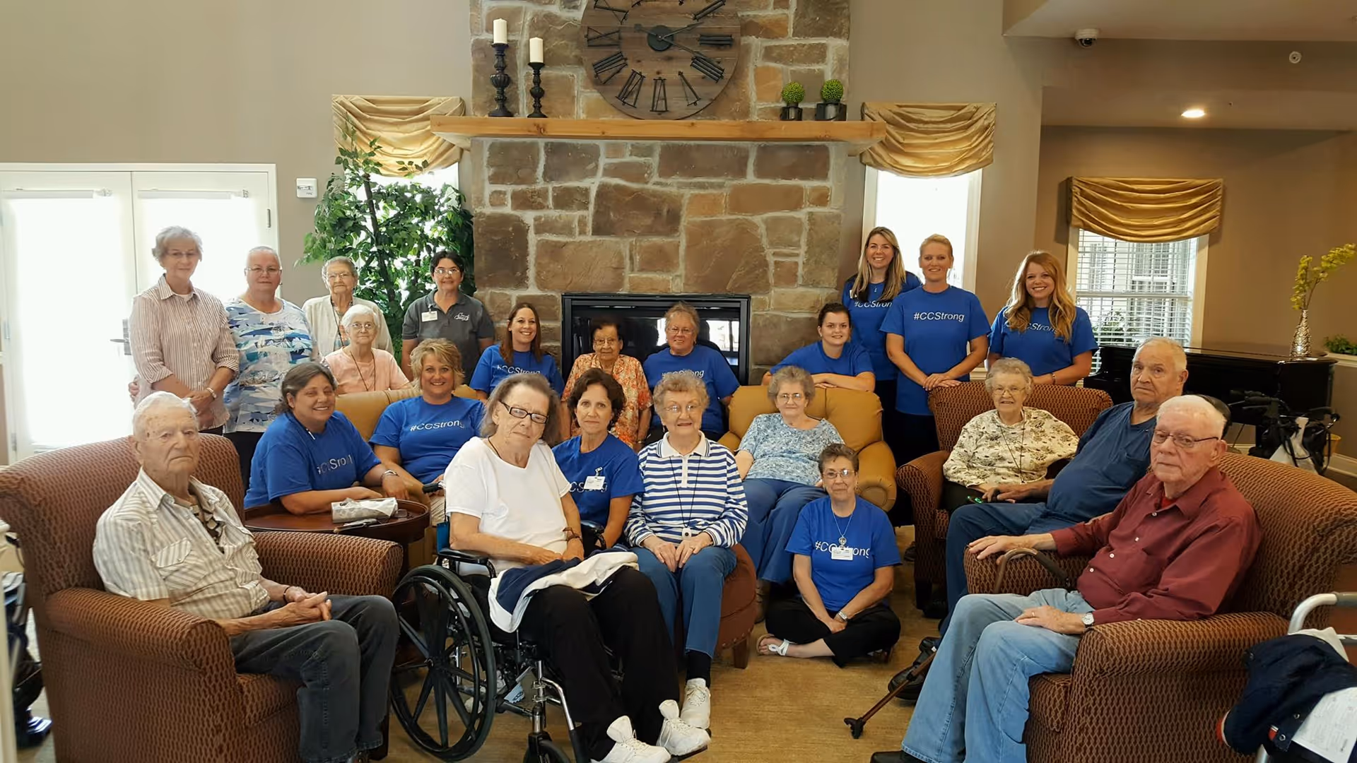 A group of senior residents and staff gathered in a lounge area with sofas and a large stone fireplace.