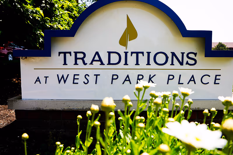 Outdoor entrance sign reading "TRADITIONS AT WEST PARK PLACE" with flowering plants in the foreground.
