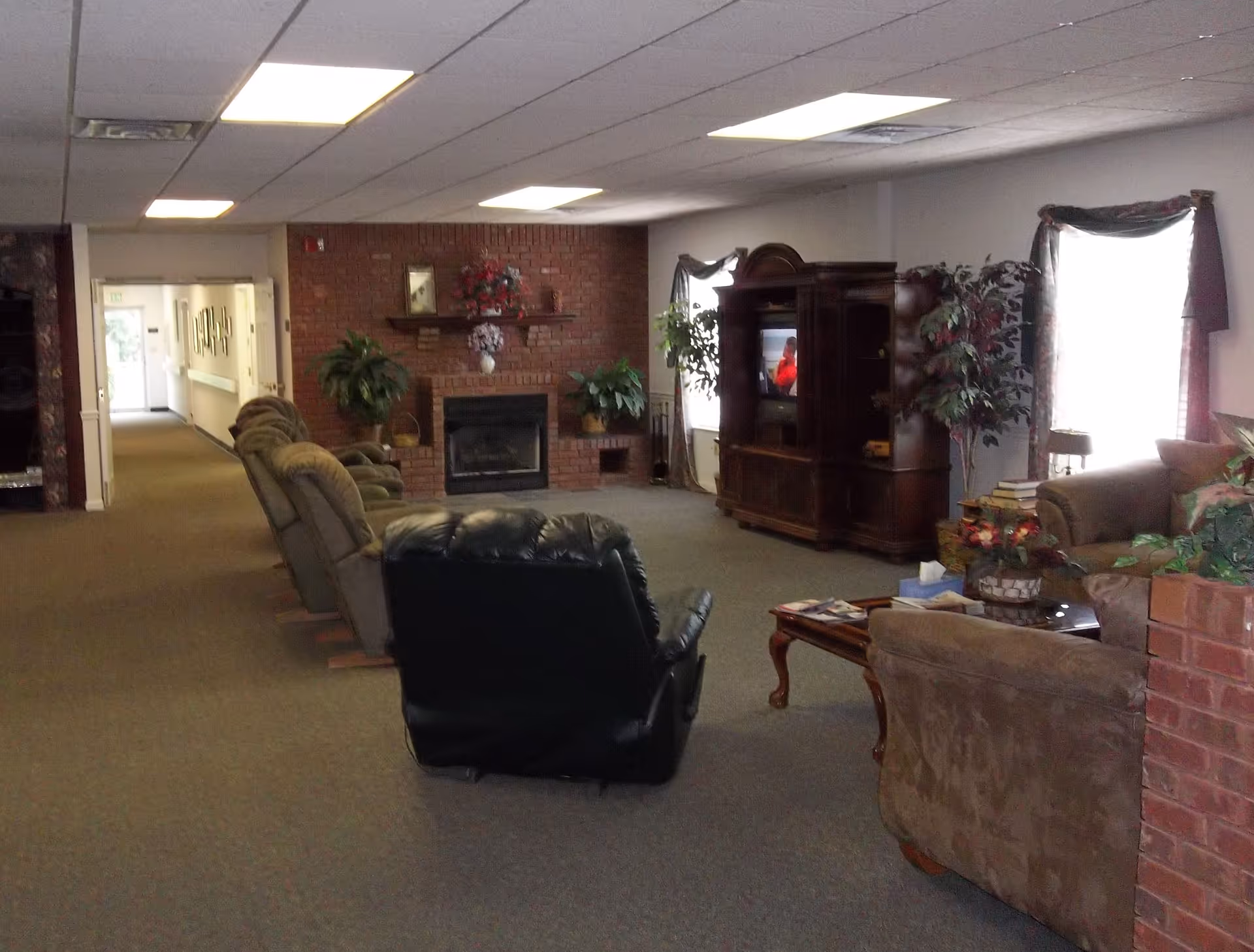 A cozy living room area in Pleasant Villa Retirement Home featuring a row of recliner chairs facing a brick fireplace with plants and floral decorations on the mantel. There is a wooden entertainment center with a TV, two windows with curtains, and additional seating including a sofa and armchair. The room is carpeted and has a hallway leading to other parts of the facility.