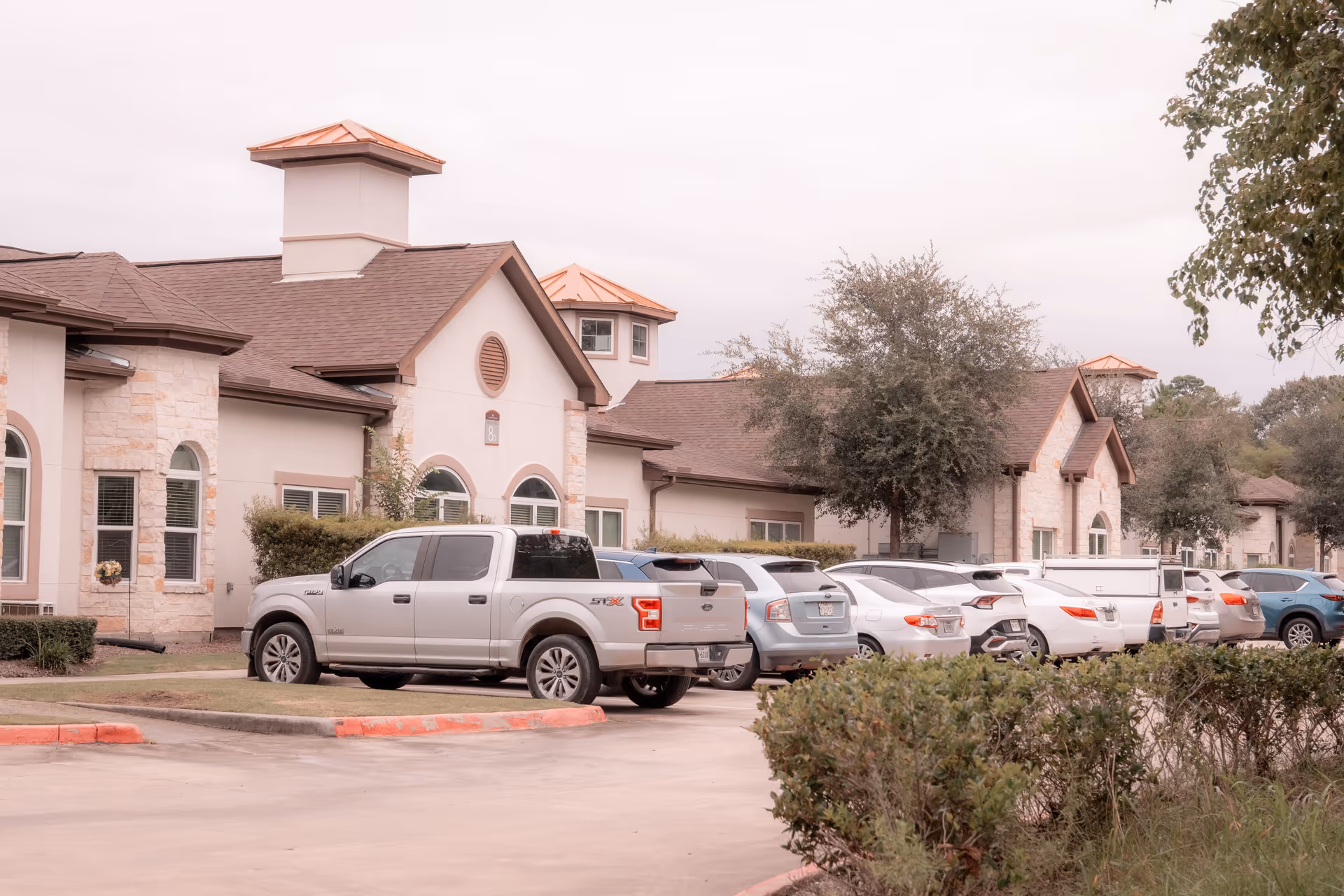 Exterior view of Woodhaven Village facility showing a parking lot with several parked cars and a pickup truck. The building has a beige stone and stucco facade with brown roofing and copper-colored cupolas. Trees and bushes are visible around the parking area.