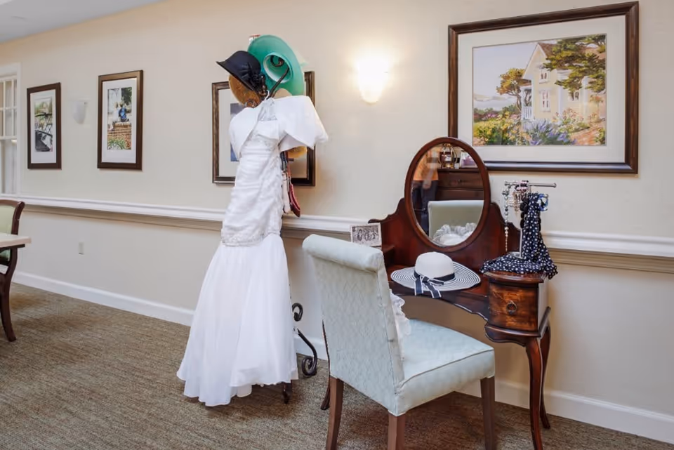 A vintage-style wooden vanity table with an oval mirror, a light blue upholstered chair, and various hats and accessories displayed on the table and a nearby dress form wearing a white dress and multiple hats. The wall behind features framed paintings and a wall sconce light.