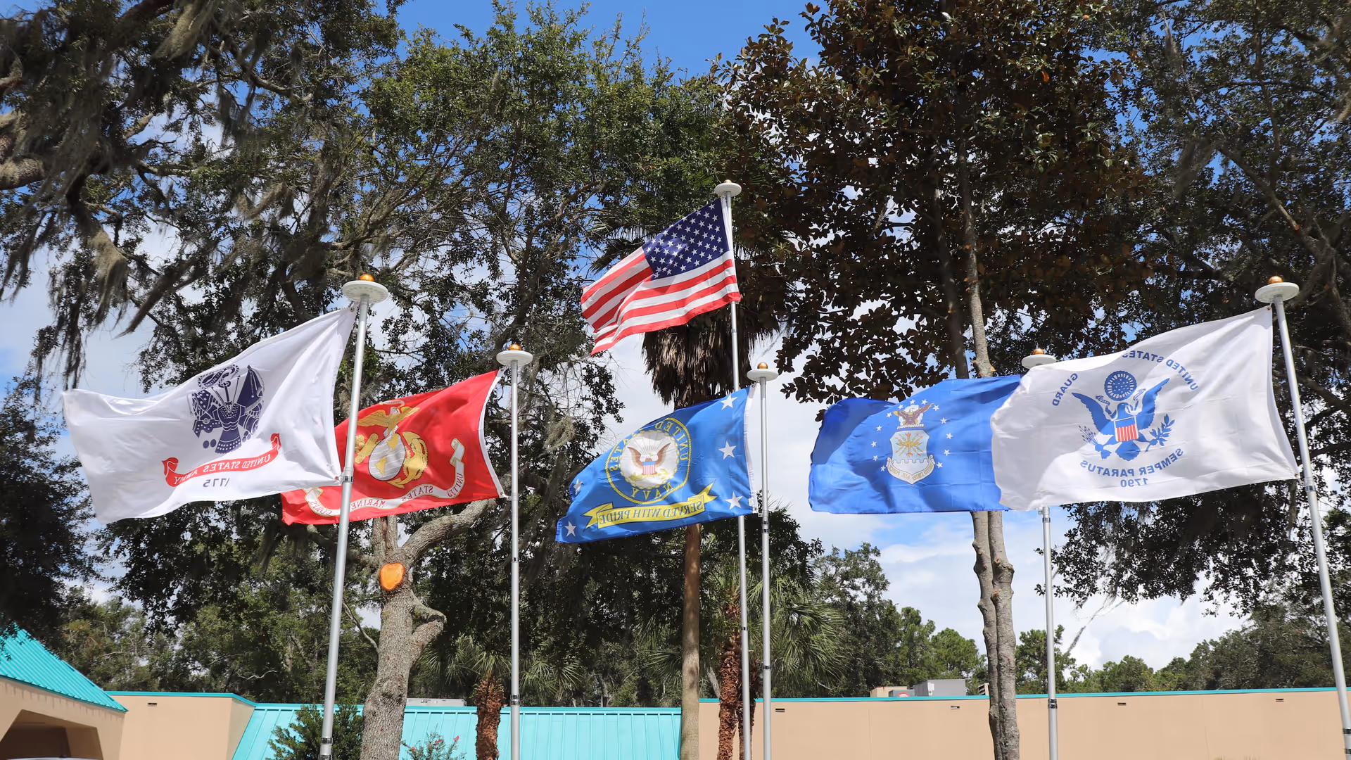Several flags, including the U.S. flag and military service flags, flying on poles in front of trees and a low building.