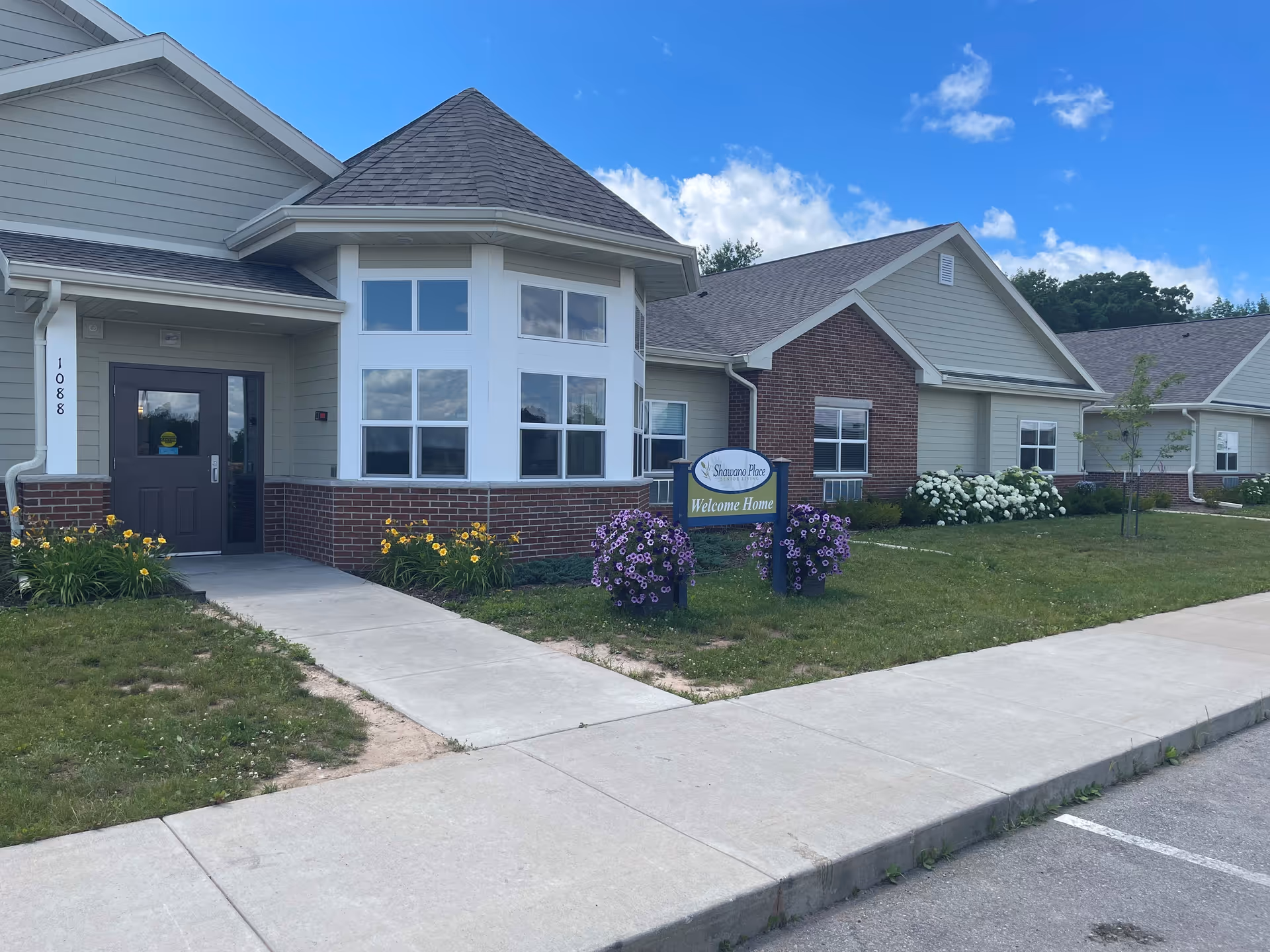 Exterior view of Shawano Place Senior Living building with a sidewalk leading to the entrance. The building features a combination of brick and siding with a large bay window and a sign that reads 'Shawano Place Welcome Home' surrounded by flowers. The sky is blue with some clouds.