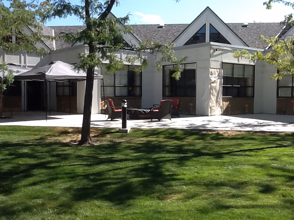 Outdoor patio area of a senior living facility with green grass, trees, a canopy tent, and several cushioned chairs arranged around a fire pit in front of a building with large windows and a sloped roof.