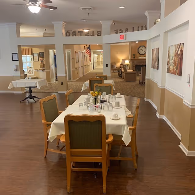 A dining area in an assisted living facility with a rectangular table covered with a white tablecloth, set with cups, glasses, and a small flower arrangement. The room has wooden floors, beige and white walls, and several chairs around the table. In the background, there is a hallway with an American flag and a cozy sitting area with armchairs and a fireplace.