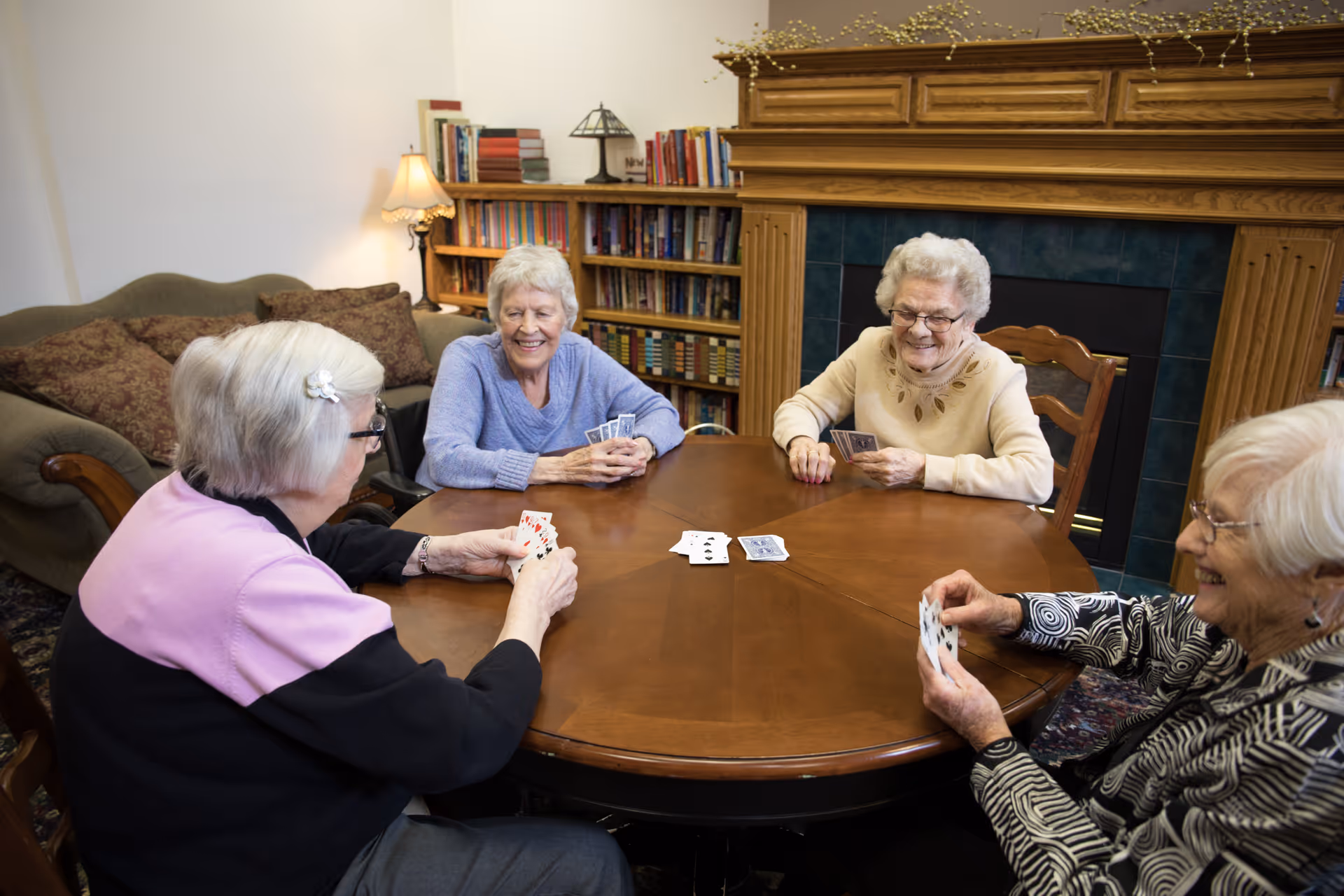 Four elderly women sitting around a wooden table playing cards in a cozy common room with a fireplace and bookshelf.