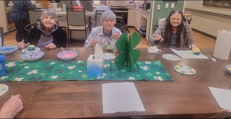 Three elderly women sitting at a wooden table in a dining area, enjoying desserts served in colorful paper bowls. The table is decorated with a green shamrock-themed runner and a green shamrock centerpiece. In the background, a kitchen area is visible with appliances and cabinets.