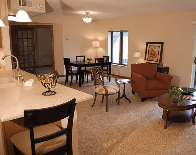 A cozy living and dining area in an independent living facility. The room features a beige carpet, a dark wooden dining table with four chairs, a red upholstered armchair, a wooden chair with a cushioned seat, and two round wooden side tables. There is a kitchen counter with a sink and a high chair in the foreground. The walls are light-colored with a framed floral artwork and two table lamps providing warm lighting. A window lets in natural light.