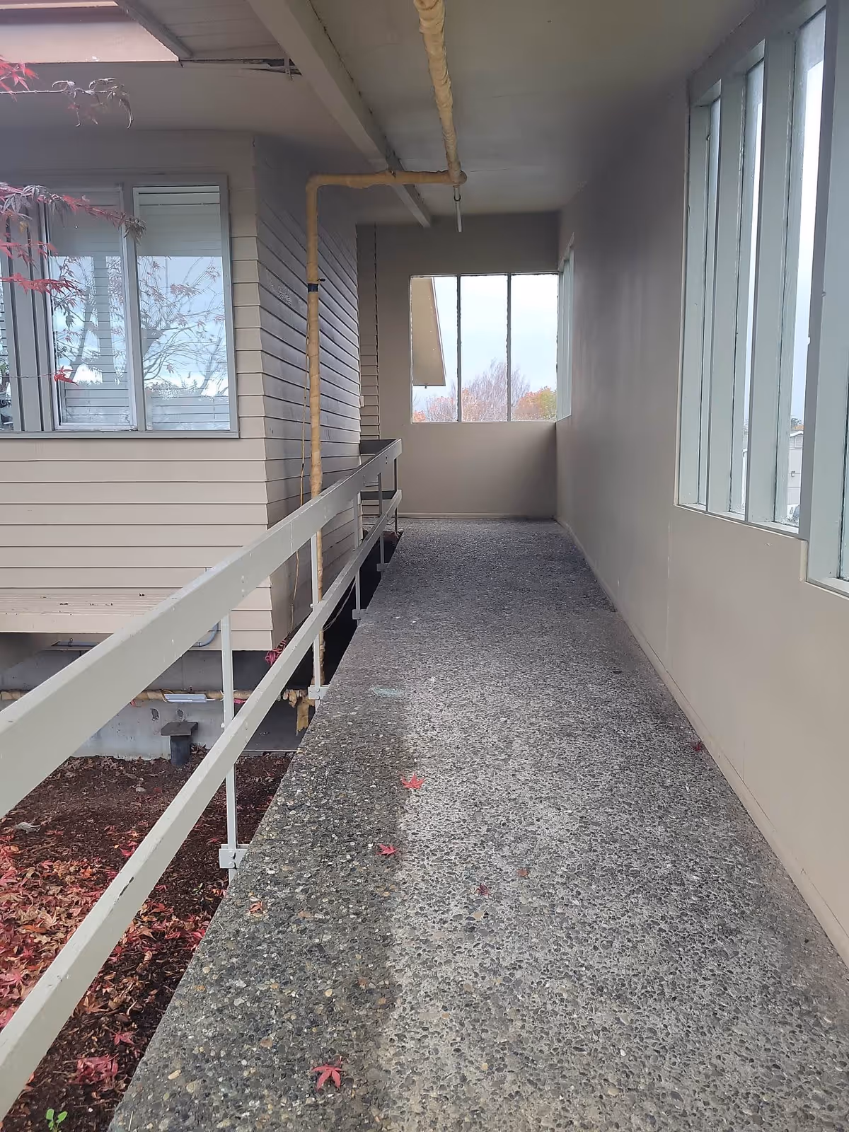 A covered outdoor walkway with a textured concrete floor and beige railings on the left side. The walkway is adjacent to a building with beige siding and windows, and there are large windows on the right side letting in natural light. Some red leaves are scattered on the ground outside the railing.