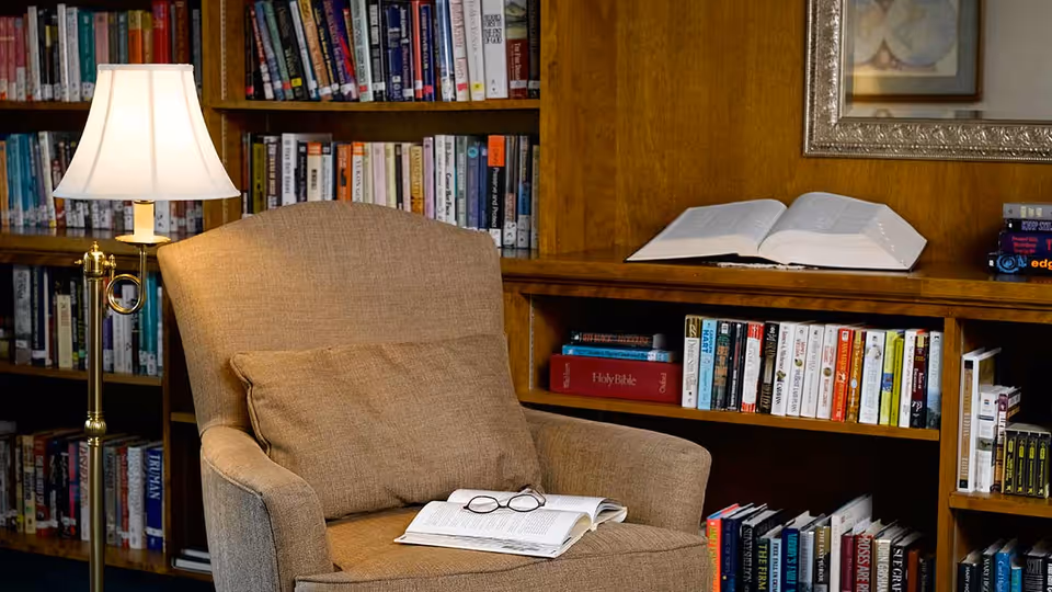 A cozy reading nook featuring a beige upholstered armchair with a matching cushion, an open book and a pair of glasses resting on the seat. Behind the chair is a wooden bookshelf filled with various books, including a prominently placed Holy Bible. A floor lamp with a white lampshade provides warm lighting to the area.