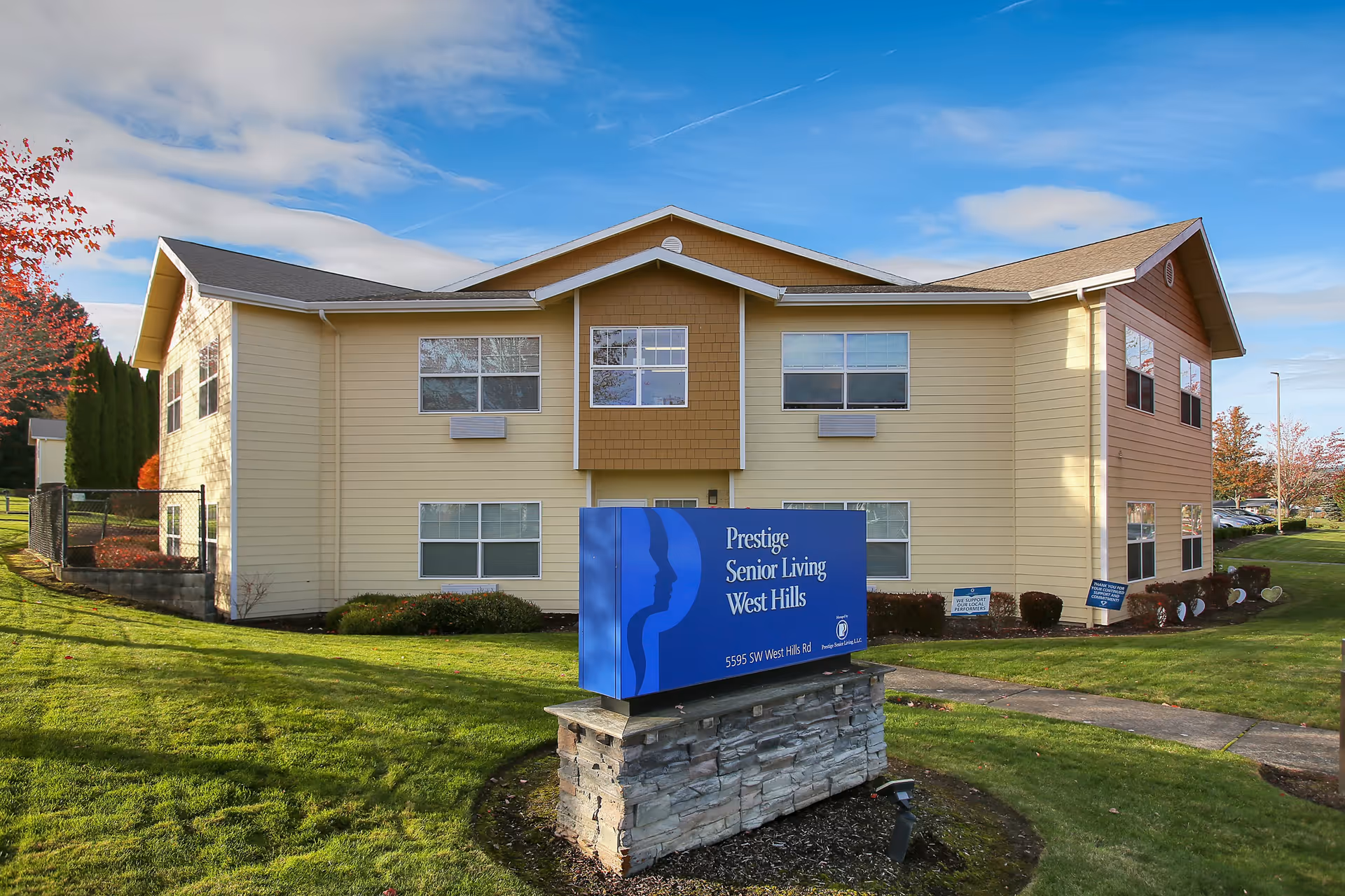 Exterior view of a two-story beige and brown building with multiple windows, surrounded by green grass and trees with autumn foliage. In front of the building is a blue sign that reads 'Prestige Senior Living West Hills' along with the address '5595 SW West Hills Rd'.