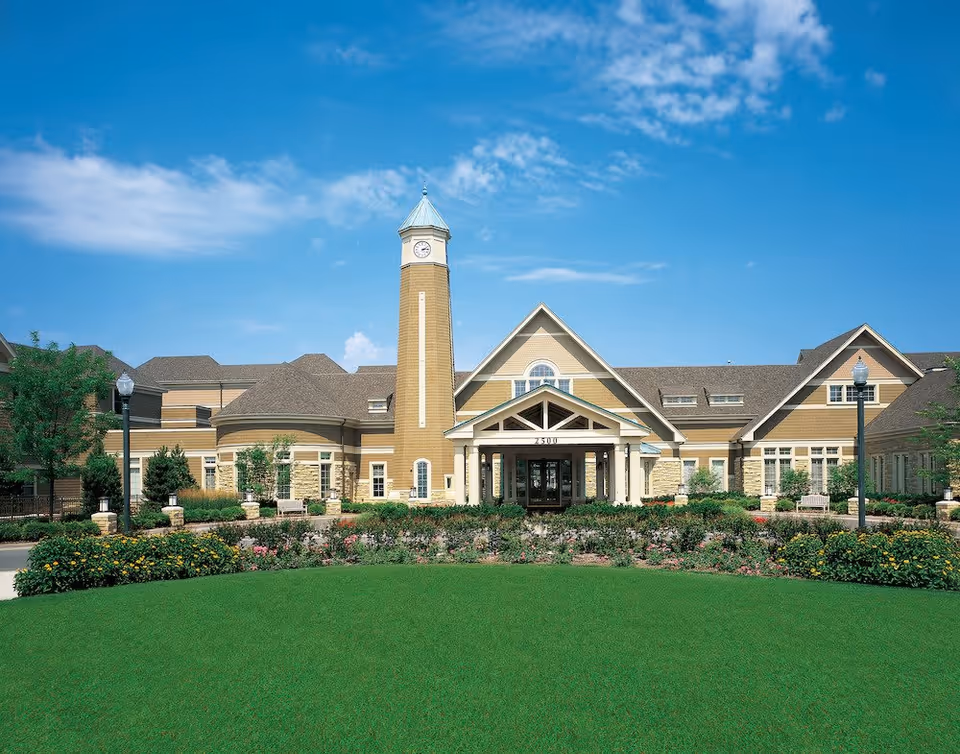 Front exterior view of a senior living facility building with a clock tower, well-maintained green lawn, flower beds, and clear blue sky.