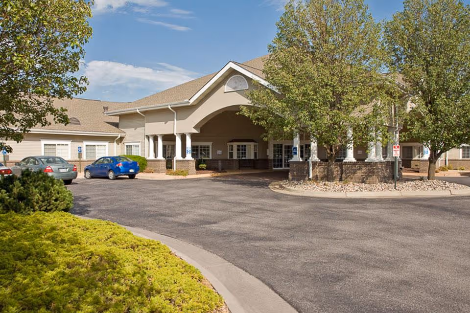 Front exterior view of Life Care Center of Wichita showing the entrance with a covered drop-off area, surrounded by trees and parked cars in the driveway under a clear blue sky.
