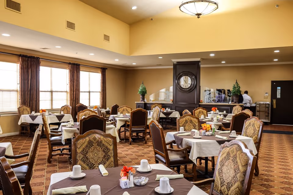 A spacious dining room with multiple tables covered in white and brown tablecloths, each set with cups, napkins, and small flower arrangements. The room features patterned upholstered chairs, large windows with brown curtains, a clock mounted on a dark wooden column, and warm beige walls. A staff member is seen near the back counter area.