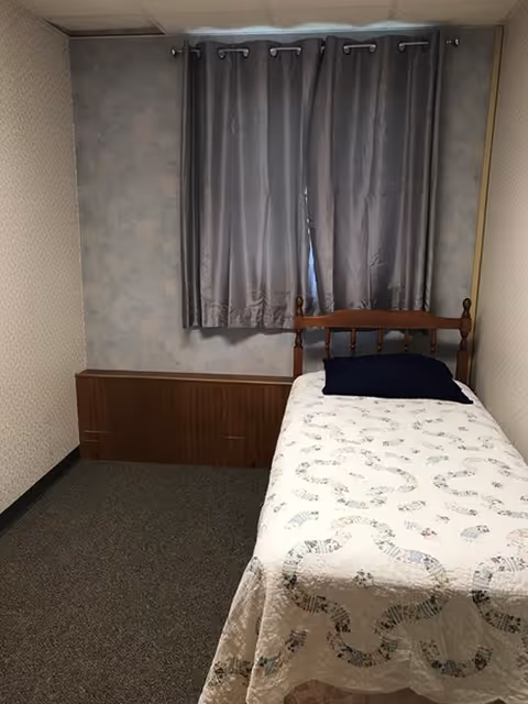 A small bedroom with a single wooden bed against the right wall. The bed is covered with a white quilt featuring a subtle pattern and has a dark pillow. Behind the bed is a window with closed gray curtains. The walls have a light patterned wallpaper, and the floor is carpeted in a dark color.