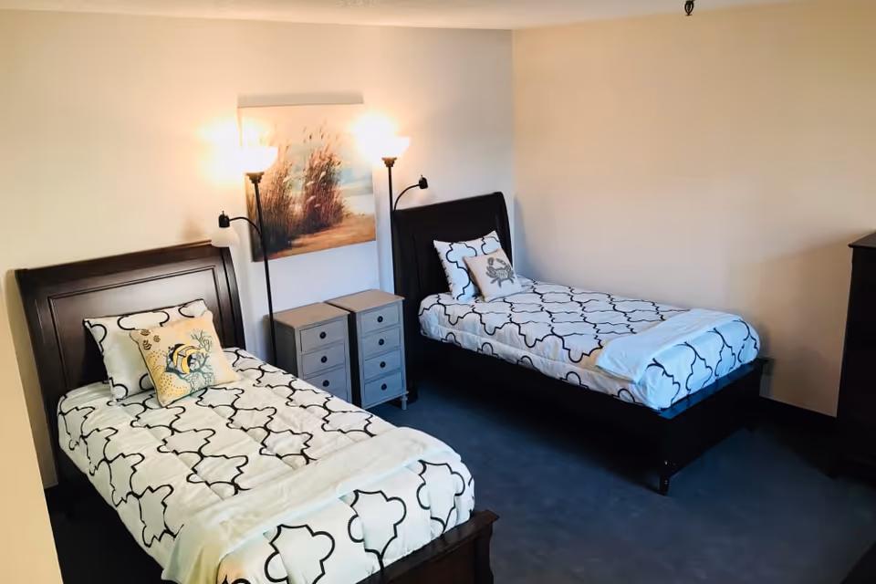 A bedroom with two twin beds featuring dark wooden frames and white bedding with black geometric patterns. Each bed has decorative pillows, and there are two small gray nightstands between the beds. Two floor lamps with white shades are positioned behind the nightstands, and a framed artwork depicting a nature scene hangs on the wall above them.