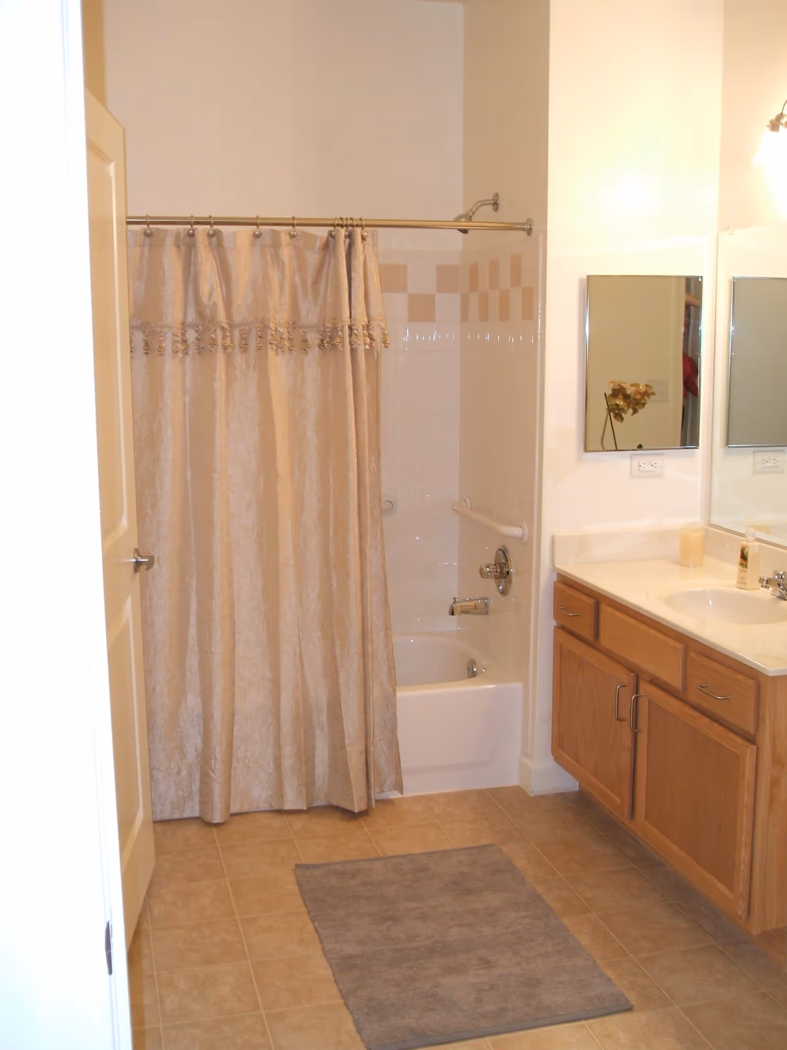 Bathroom with a beige shower curtain covering a bathtub, beige tiled floor with a gray bath mat, wooden vanity with a white countertop and sink, two mirrors above the sink, and a wall-mounted light fixture.