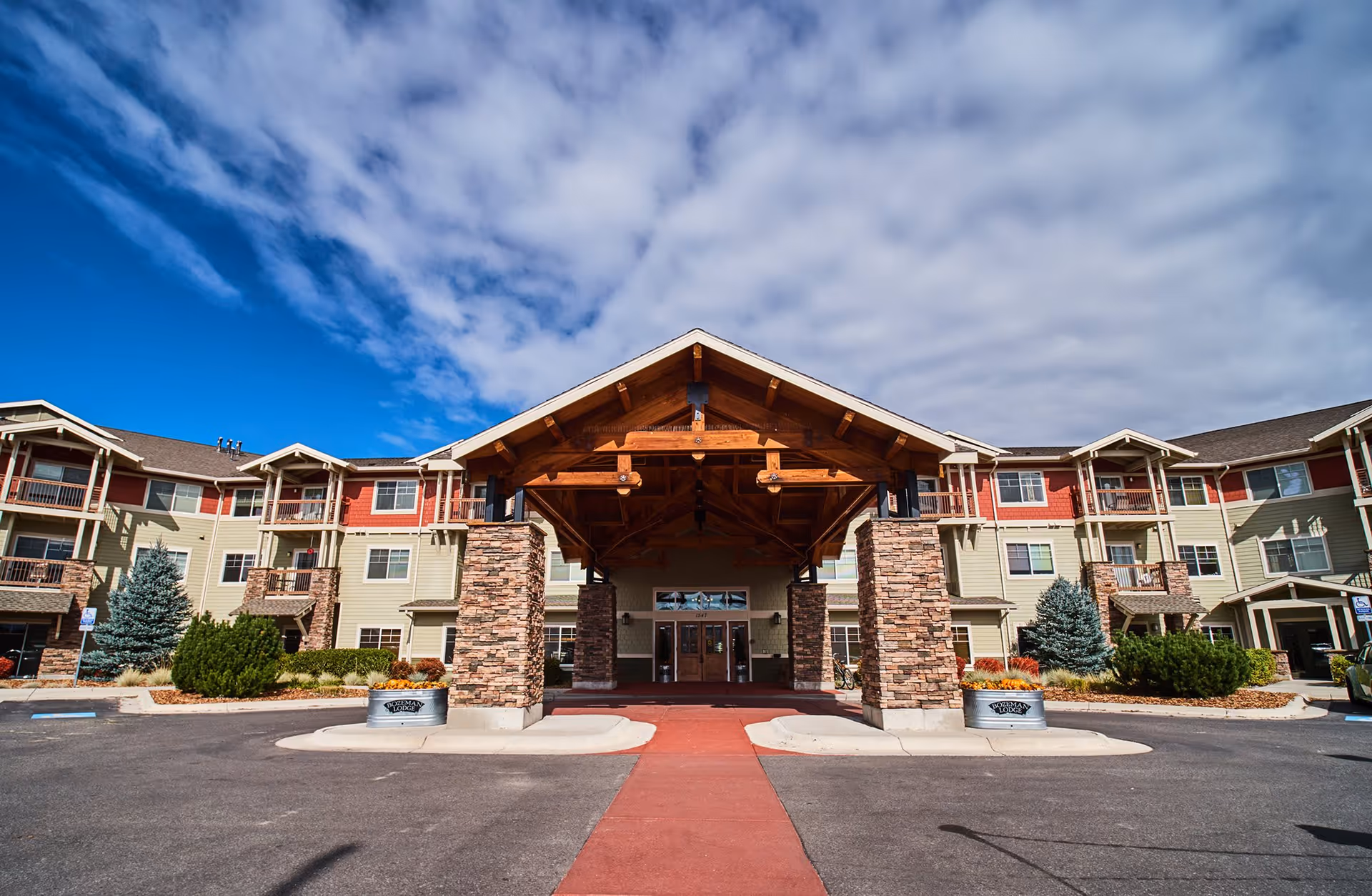 Front exterior view of Bozeman Lodge, a multi-story building with a large covered entrance supported by stone pillars, surrounded by landscaped bushes and trees under a partly cloudy blue sky.