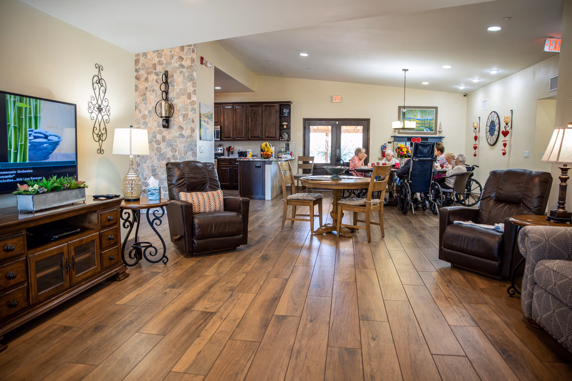 A spacious assisted living common area with wooden flooring, two brown recliner chairs, a wooden TV stand with a flat-screen TV, and a round wooden table with chairs. In the background, several elderly residents are seated around a table near the kitchen area, engaged in conversation. The room is warmly lit with lamps and ceiling lights, and decorated with wall art and a clock.