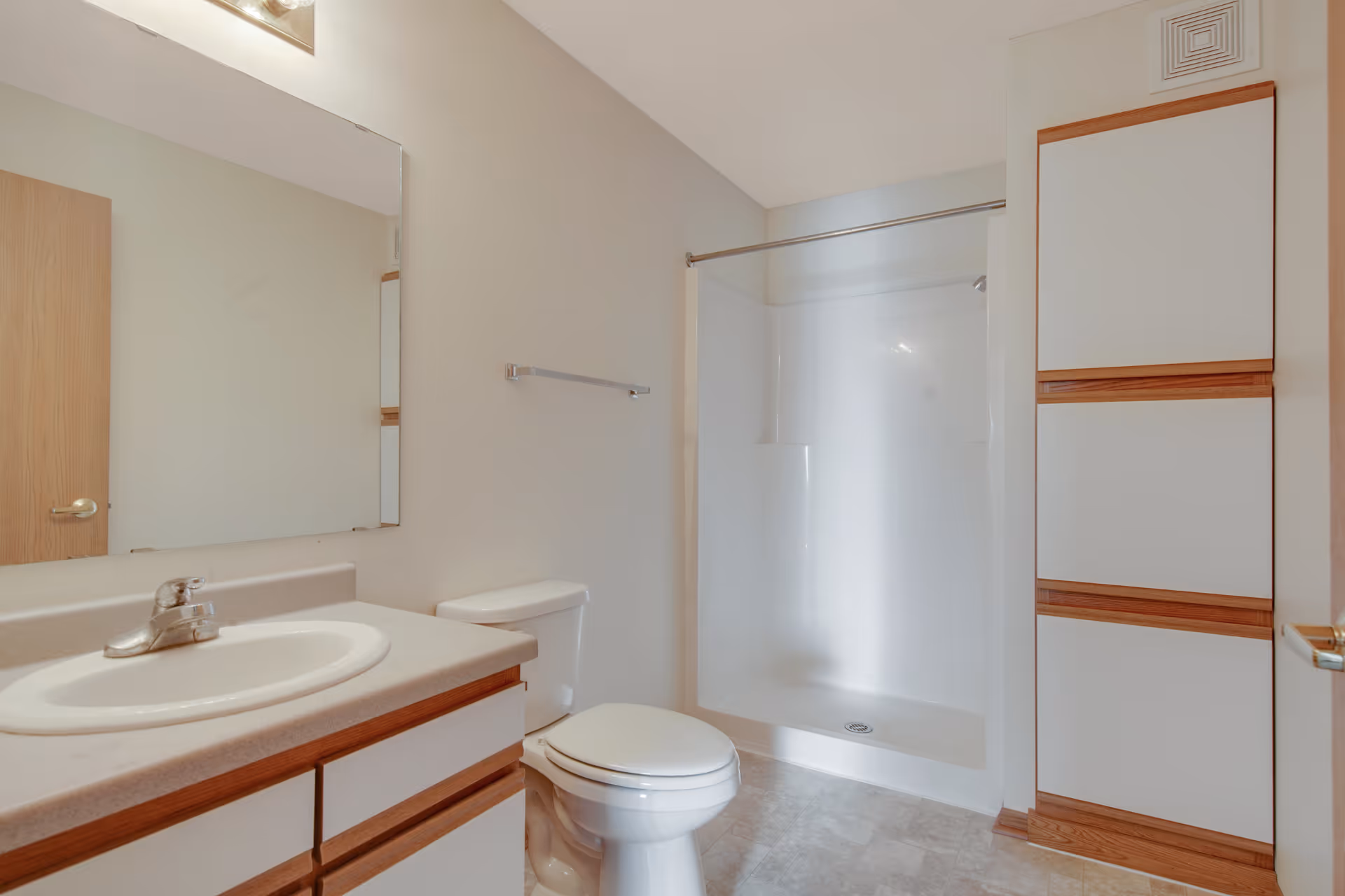 A clean bathroom featuring a sink with a countertop and cabinet, a toilet, a walk-in shower with a curtain rod, and a tall storage cabinet with white doors and wooden trim. The walls are light-colored, and there is a large mirror above the sink.