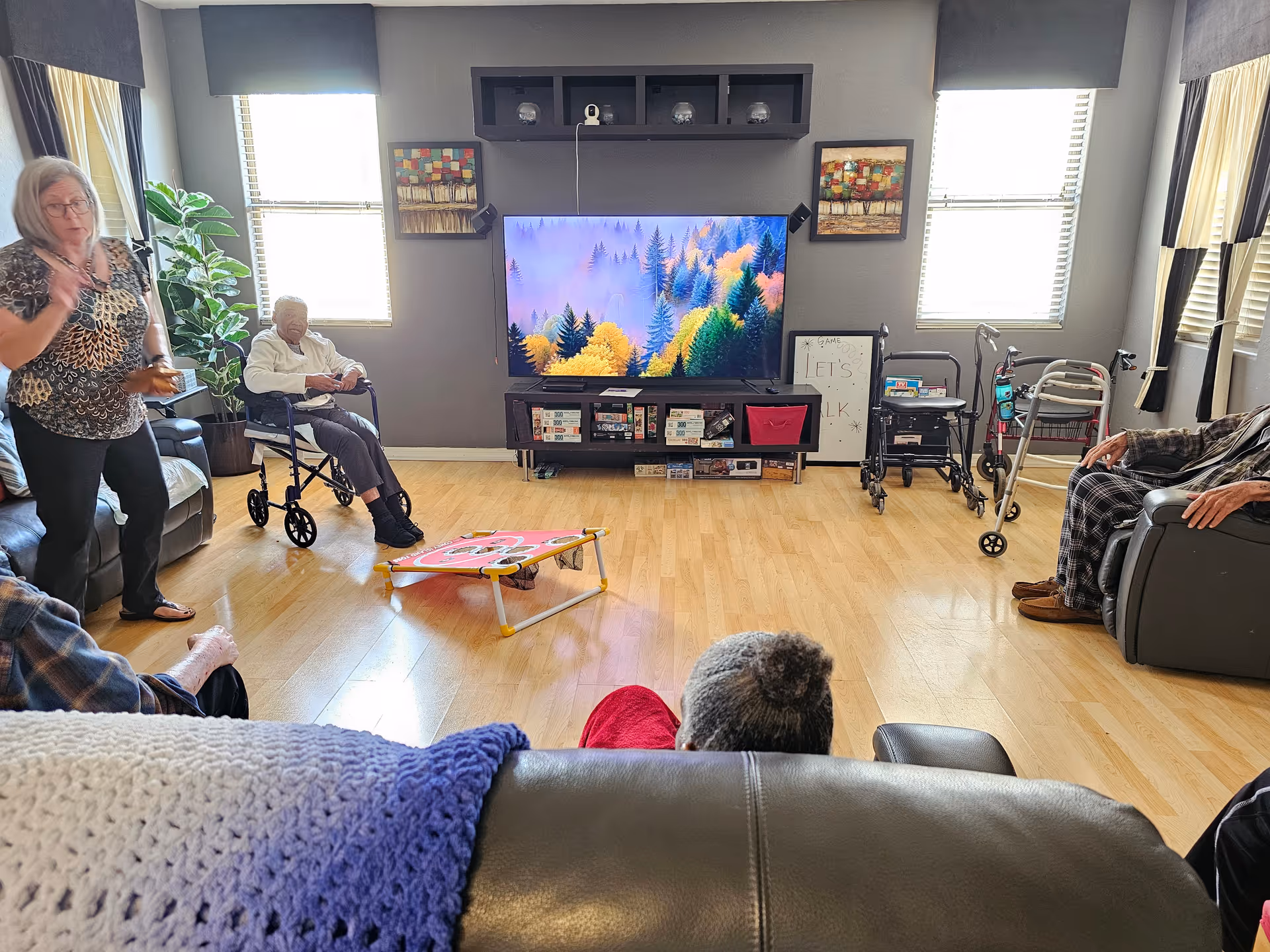 A senior living facility common room with several elderly residents seated in chairs and wheelchairs around a large flat-screen TV mounted on a gray wall. The room has wooden flooring, two windows with blinds, a potted plant, and various mobility aids such as walkers. A woman stands on the left side of the room, and a cornhole game board is on the floor in the center.