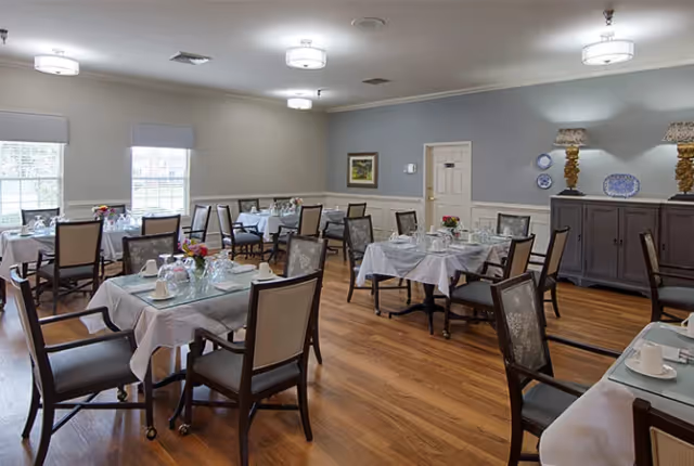 A dining room with several tables covered in white tablecloths, each set with plates, glasses, and floral centerpieces. The room has wooden floors, light gray walls with white trim, and multiple windows letting in natural light. There are cushioned chairs around the tables and decorative lamps and plates on a sideboard against the wall.