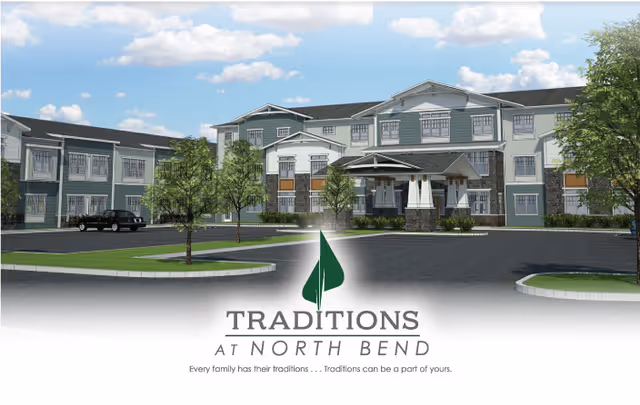 Exterior view of Traditions at North Bend, a large three-story senior living facility with a covered entrance, surrounded by trees and a parking area under a partly cloudy sky.