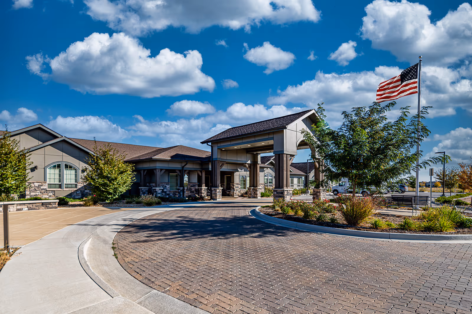 Exterior view of Aspens at Fort Collins Memory Care facility showing the entrance with a covered driveway, landscaped greenery, an American flag on a flagpole, and a partly cloudy blue sky.