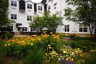 Outdoor garden area with blooming yellow and purple flowers, green shrubs, and trees in front of a white multi-story senior living facility building with multiple windows and a patio area with tables and chairs.
