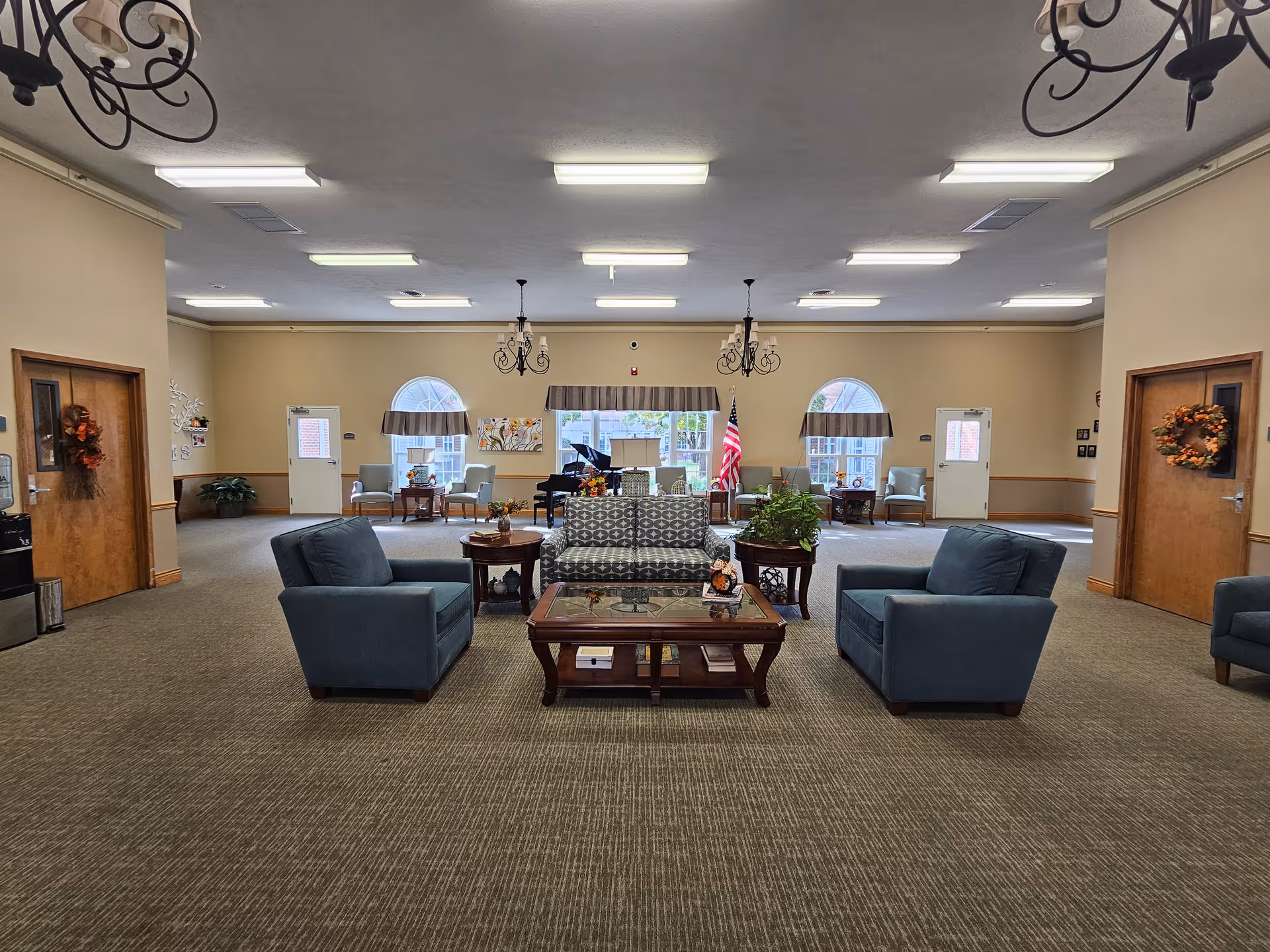 Spacious common lounge with sofas and armchairs arranged around a central coffee table, chandeliers, a piano and windows in the background.