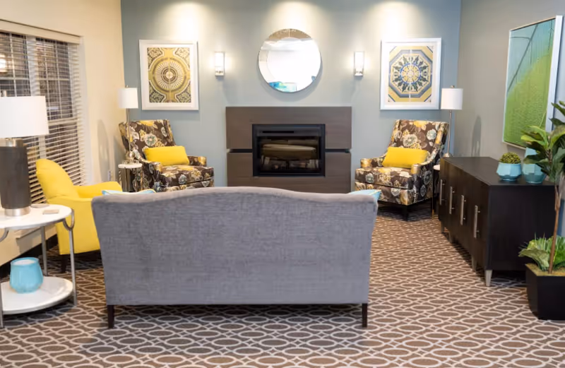 A cozy common living area with a sofa facing a fireplace, patterned armchairs, side tables and decorative accents.