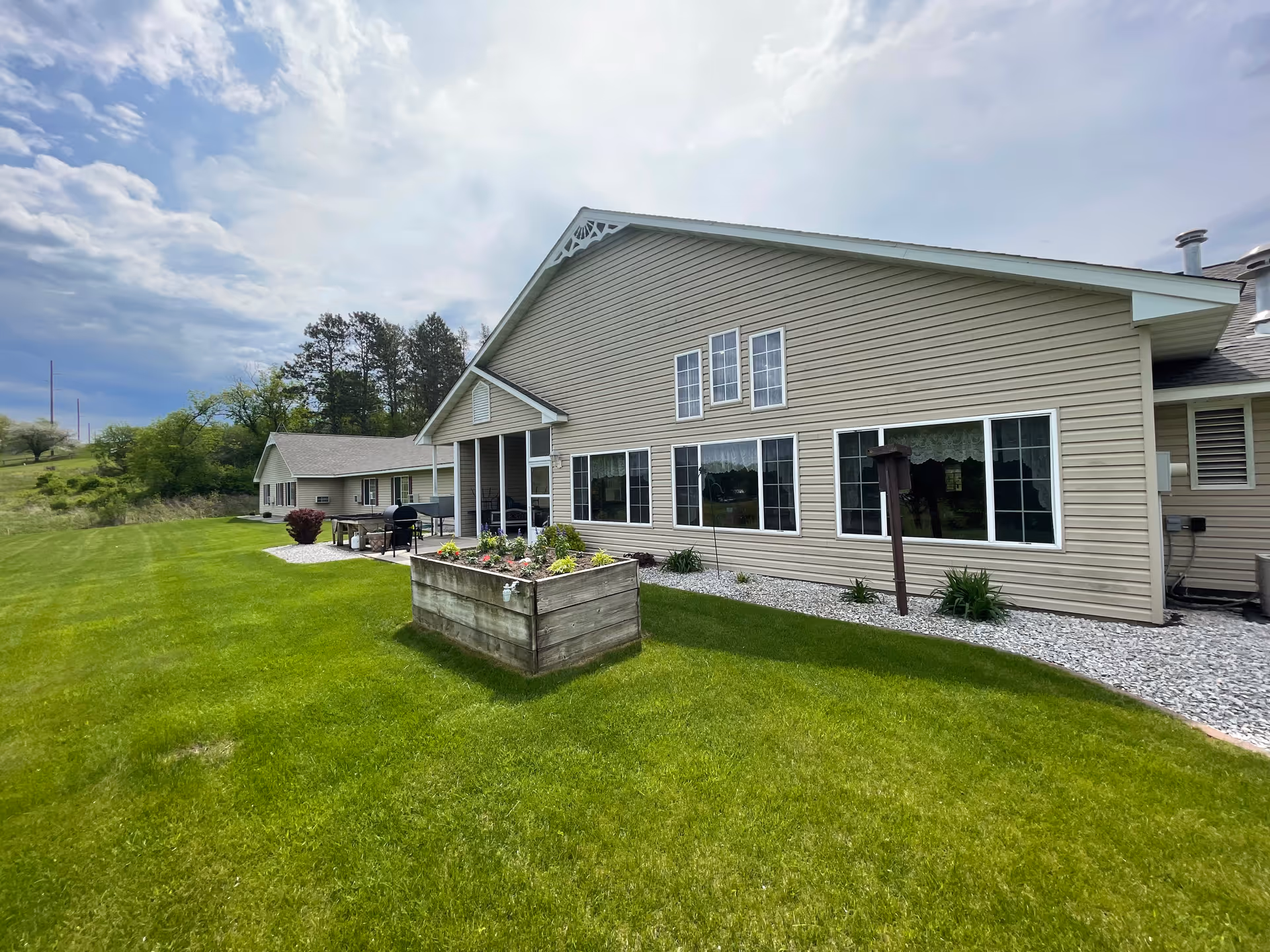 Exterior view of a beige single-story building with multiple windows, a small covered porch area, and a raised garden bed in front. The building is surrounded by a well-maintained green lawn and some trees in the background under a partly cloudy sky.