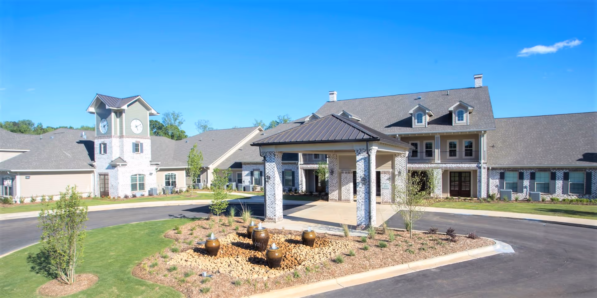 Exterior view of The Claiborne at McComb senior living facility showing a large building with a clock tower, covered entrance, landscaped garden with decorative pots, and a circular driveway under a clear blue sky.
