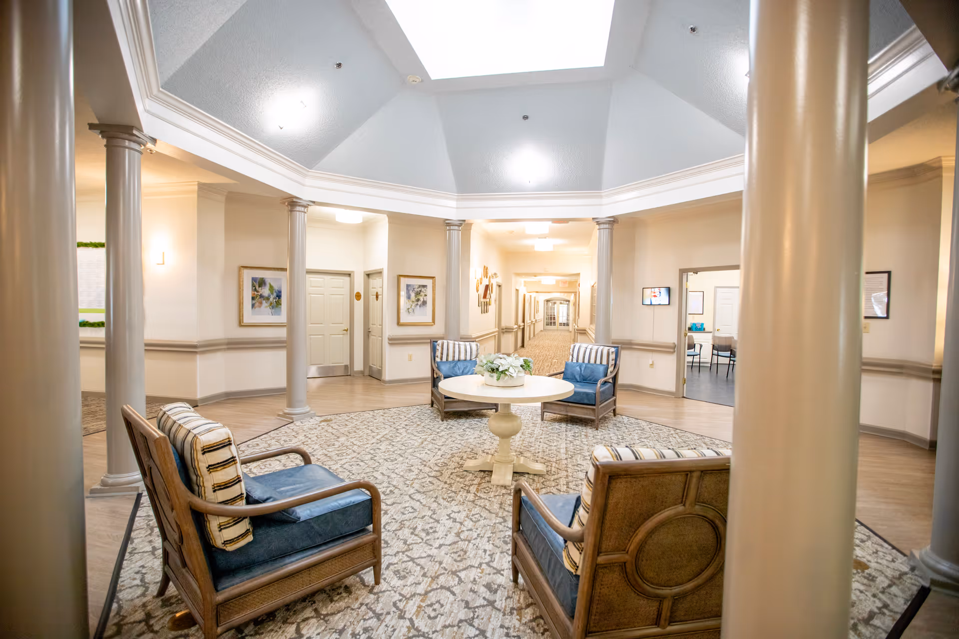 A spacious, well-lit common area in a senior living facility featuring four wooden armchairs with blue cushions and striped pillows arranged around a round white table with a floral centerpiece. The room has a high ceiling with recessed lighting, beige walls, decorative columns, and a patterned carpet. Doors and framed artwork are visible along the walls, with a hallway extending in the background.