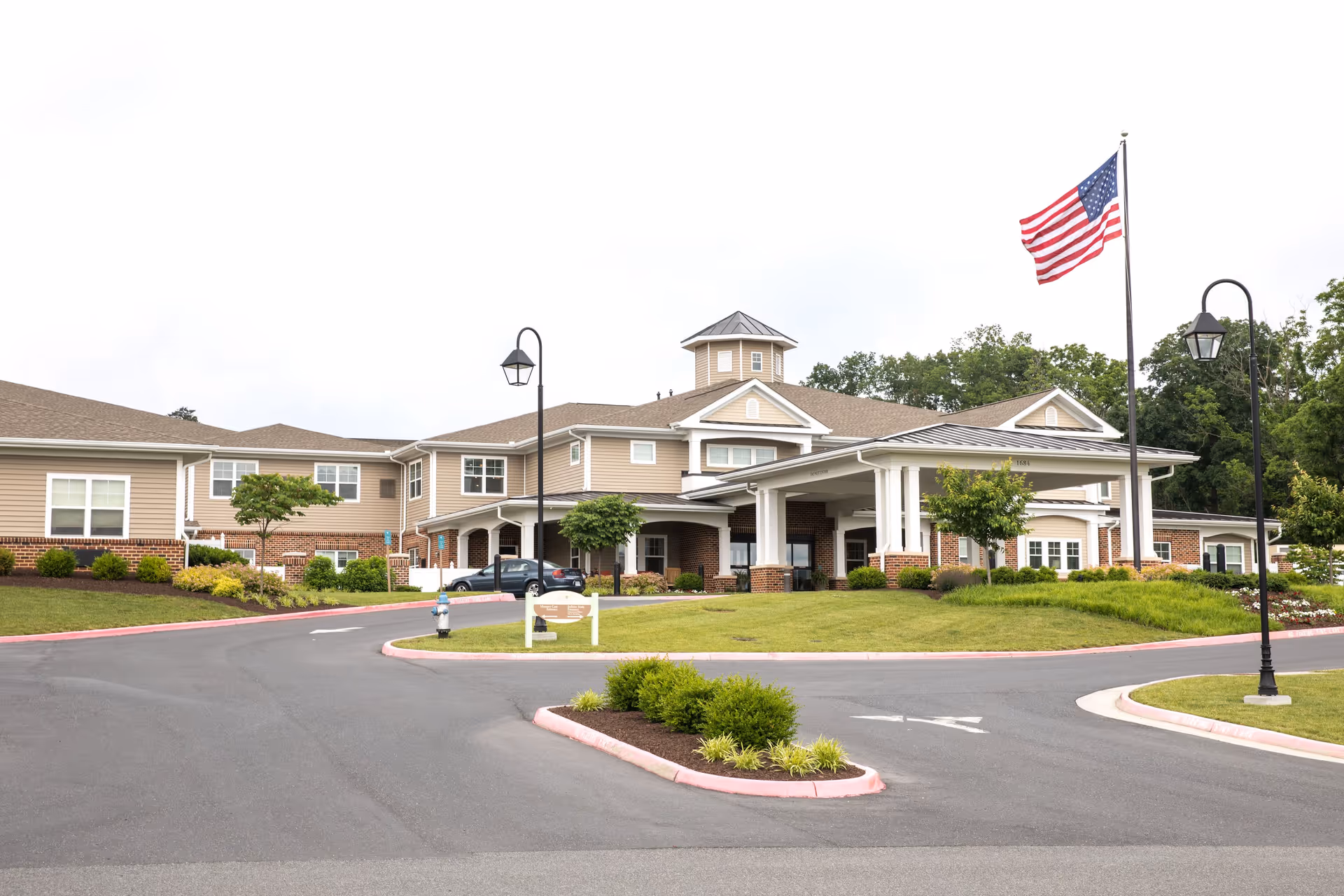 Exterior view of Bellaire at Stone Port Assisted Living & Memory Care facility showing a large building with beige siding and brick accents, a covered entrance, landscaped greenery, an American flag on a tall flagpole, and street lamps along the driveway.