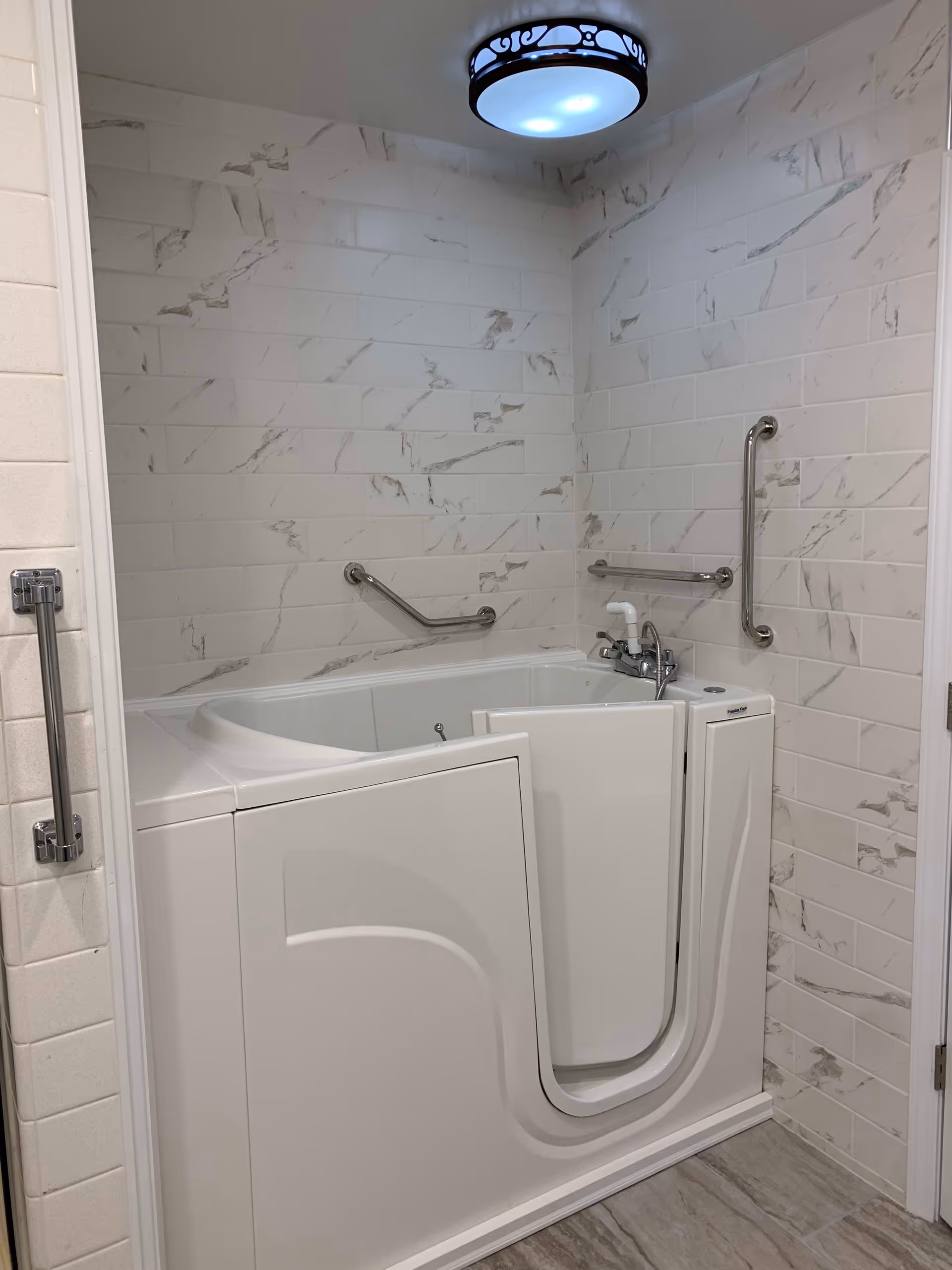 A walk-in bathtub with an open door, surrounded by white marble-patterned tiled walls. The bathtub has multiple grab bars for safety and a faucet with a handheld showerhead. The floor is tiled in a light gray color, and a round ceiling light fixture is visible above.