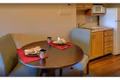 A small dining area with a round wooden table set for two with red placemats, napkins, and coffee mugs. Two striped upholstered chairs are positioned at the table. In the background, there is a compact kitchenette with wooden cabinets, a microwave, a small refrigerator, and a sink.