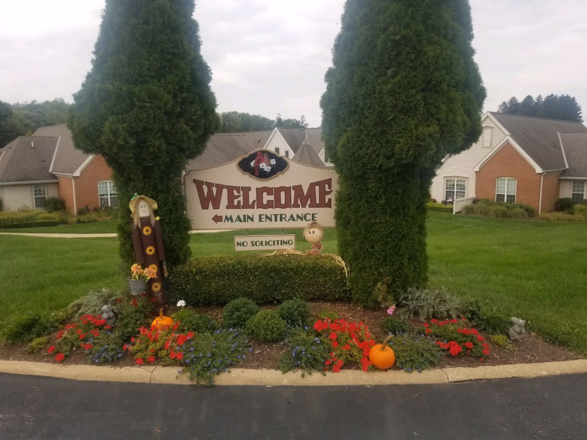 A landscaped entrance area with a large welcome sign reading 'WELCOME MAIN ENTRANCE' and 'NO SOLICITING' beneath it, flanked by two tall evergreen trees. The area is decorated with colorful flowers, pumpkins, and small scarecrow figures. In the background, there are residential-style buildings with brick and siding exteriors and well-maintained lawns.